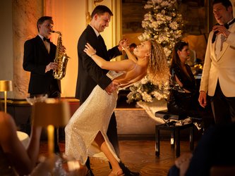en:A woman in a white dress and a man in a black suit dancing with musicians in the background; fr:Une femme en robe blanche et un homme en costume noir qui danse avec des musiciens en fond