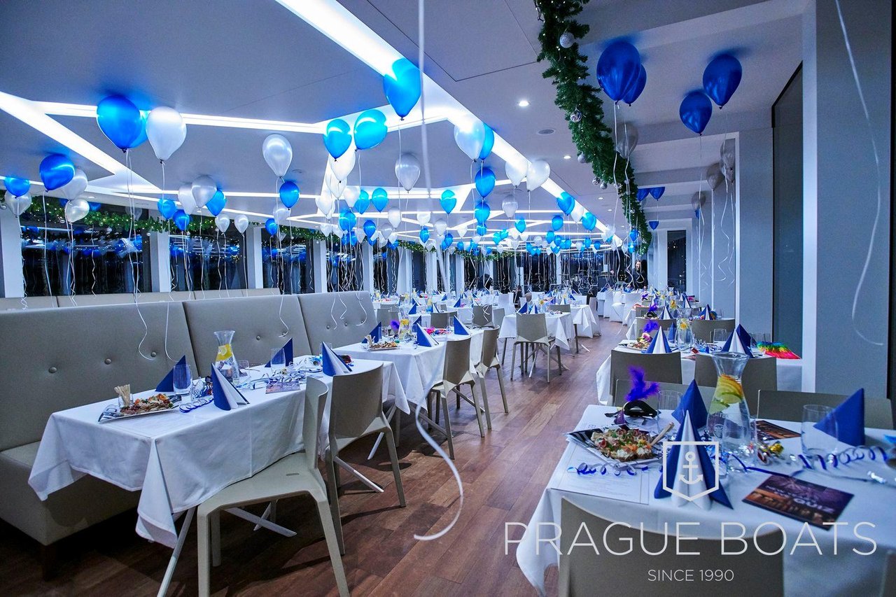 Several tables with gray armchairs, the party is taking place on a boat, and there are blue and white balloons