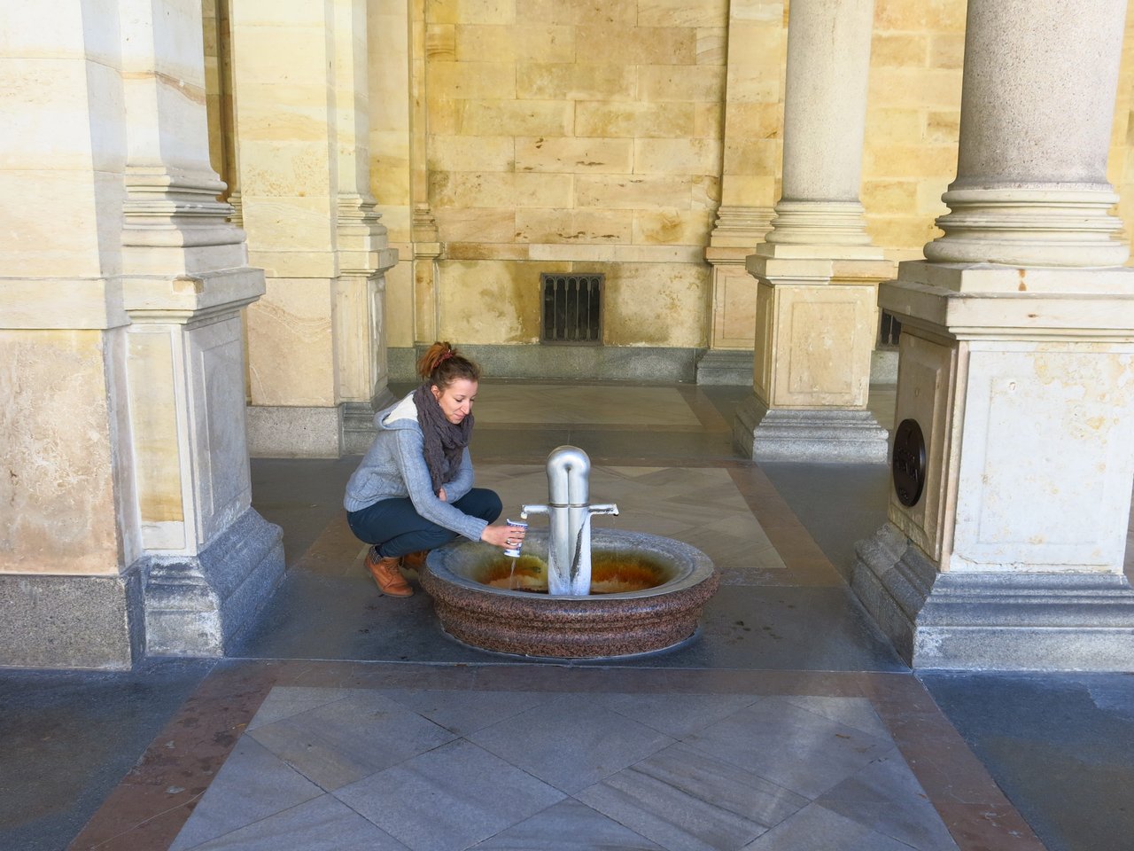 A person filling a glass at a small fountain surrounded by stone pillars
