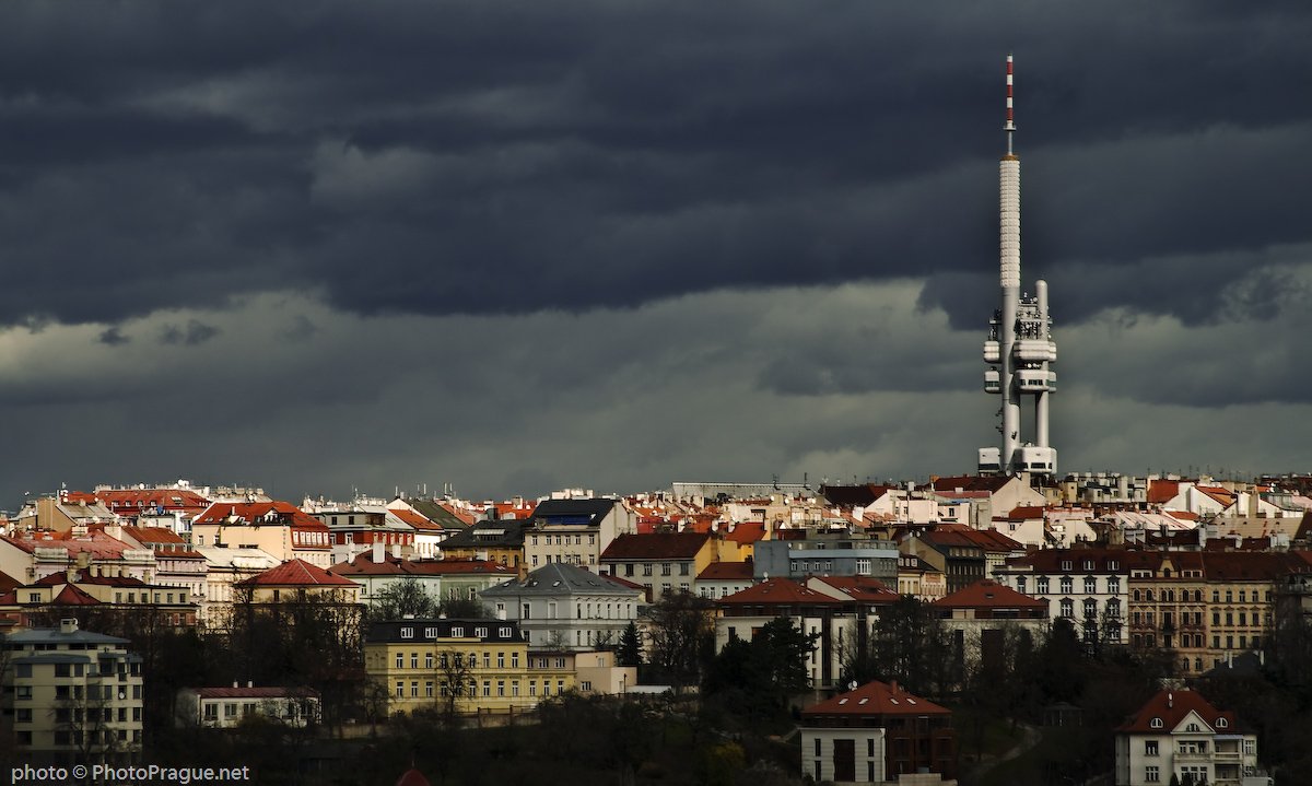 A tall, pointed white tower rises above a multitude of houses against a very dark sky filled with gray clouds.